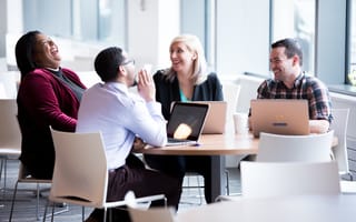 Photo of four colleagues sitting at a table with laptops open, laughing at a joke.
